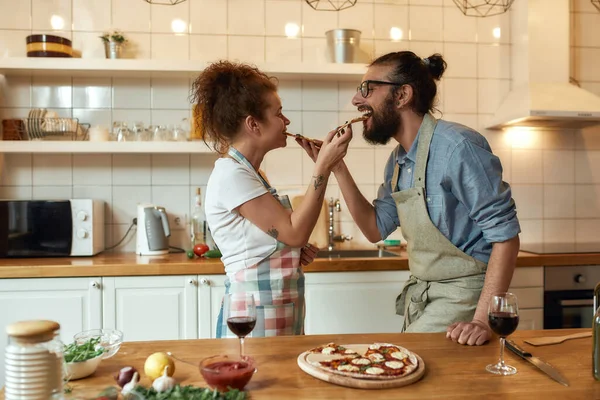 Assaggialo. Giovane uomo e donna in grembiule che si nutrono la pizza appena sfornata mentre sono in cucina. Amore, concetto di relazioni — Foto Stock