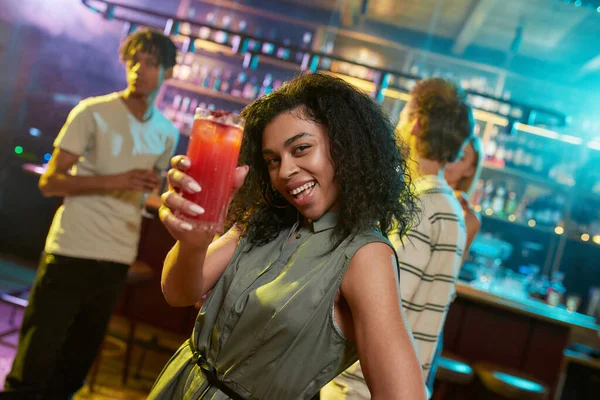 Atractiva mujer joven mestiza mirando la cámara mientras posa con un cóctel en la mano y amigos charlando, tomando bebidas en el mostrador del bar en el fondo — Foto de Stock