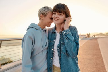 Lezbiyen çift kameraya poz veriyor, köprüde duruyor ve güneşin doğuşunu izlerken birbirlerine sarılıyorlar. Homoseksüellik, LGBT ve aşk konsepti