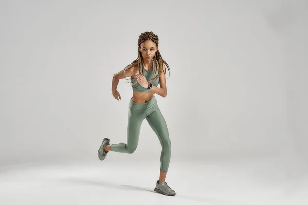멈추지 마. 활발히 달리거나 운동을 하는 젊은 산발적 인 혼혈 여성이 회색 배경 위에서 고립 된 채로 달리거나 운동을 하는 동안 카메라를 보고 있는 모습이 전체적으로 촬영되었다 — 스톡 사진
