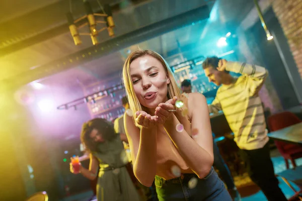 Baile y fiesta. Atractiva mujer joven caucásica soplando confeti a la cámara mientras posa en el club nocturno. Amigos bailando, celebrando, divirtiéndose en el fondo — Foto de Stock