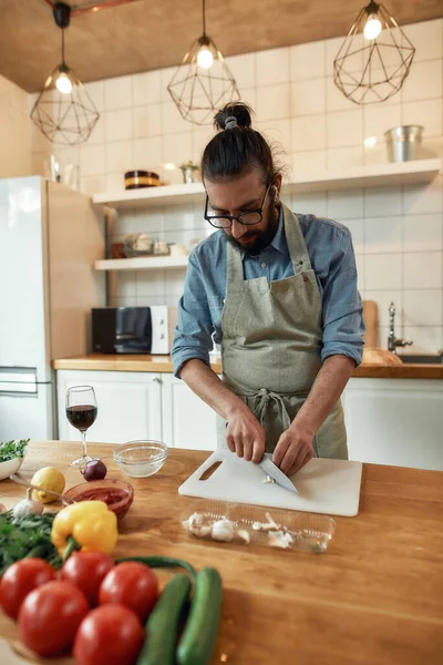 Önlüklü genç adam, yemek hazırlarken sarımsak kesiyor, mutfakta dikiliyor. Ev konseptinde aşçılık