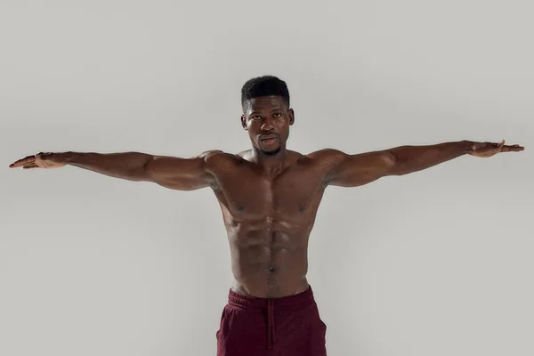Jovem homem americano africano muscular com tronco nu olhando para a câmera enquanto posando com os braços estendidos isolados sobre fundo cinza. Esportes, treino, conceito de musculação — Fotografia de Stock