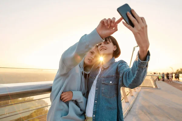 Ζευγάρι λεσβιών στέκεται στη γέφυρα, ποζάροντας βγάζοντας μια selfie φωτογραφία, βλέποντας την ανατολή του ηλίου μαζί — Φωτογραφία Αρχείου