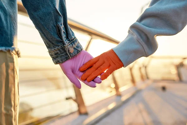 Primer plano de las manos de una pareja de lesbianas. Dos chicas sosteniendo sus manos, usando guantes protectores, de pie en el puente, admirando el amanecer juntas —  Fotos de Stock