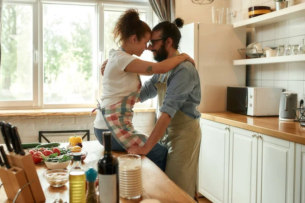 台所に立っている間、若い男は彼女を見て、抱きついていた。室内で野菜とピザを作っているカップル。料理、趣味、ライフスタイル — ストック写真