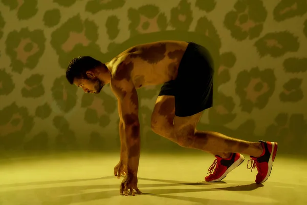 Run like a devil. Side view of a young muscular caucasian man standing in start position, ready for run isolated over yellow cheetah background
