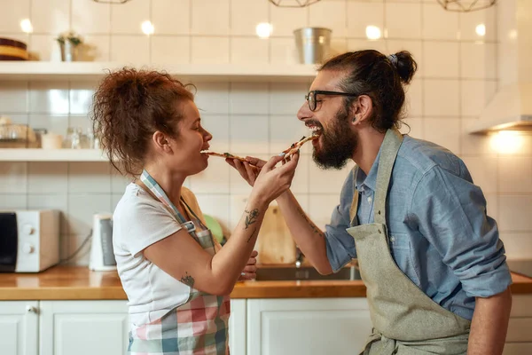 Goditi il sapore. Giovane uomo e donna in grembiule che si nutrono la pizza appena sfornata mentre sono in cucina. Amore, concetto di relazioni — Foto Stock