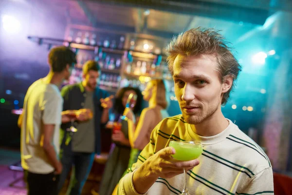 Potret pemuda kaukasia melihat kamera sambil berpose dengan koktail di tangannya dan teman-teman mengobrol, memiliki minuman di meja bar di latar belakang — Stok Foto