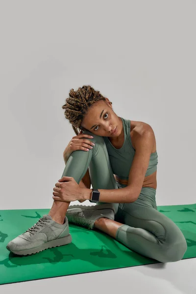 Mujer joven de raza mixta deportiva en ropa deportiva mirando a la cámara, sentada en una alfombra aislada sobre un fondo gris. Estilo de vida saludable y concepto deportivo — Foto de Stock