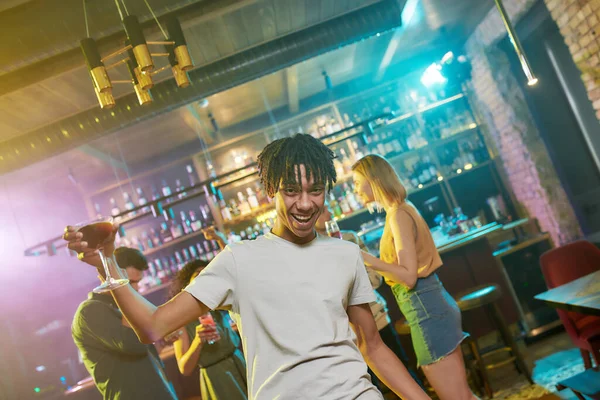Glada blandras ung man att bli full, ler mot kameran med en cocktail i handen, vänner chatta, dansa, ha kul i bakgrunden — Stockfoto