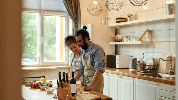 Giovane coppia innamorata di preparare l'impasto per fare la pizza con le verdure a casa. Uomo e donna indossano il grembiule, cucinano insieme in casa. Hobby, stile di vita — Foto Stock