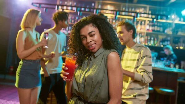 Attraktiv blandras ung kvinna tittar lekfull på kameran samtidigt poserar med en cocktail i handen. Vänner chattar, dricka vid bardisken i bakgrunden — Stockfoto