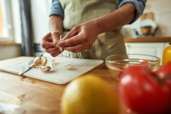 Närbild av händerna på mannen i förkläde skalning vitlök samtidigt förbereda hälsosam måltid, soppa i köket. Matlagning hemma, italienskt kök koncept — Stockfoto