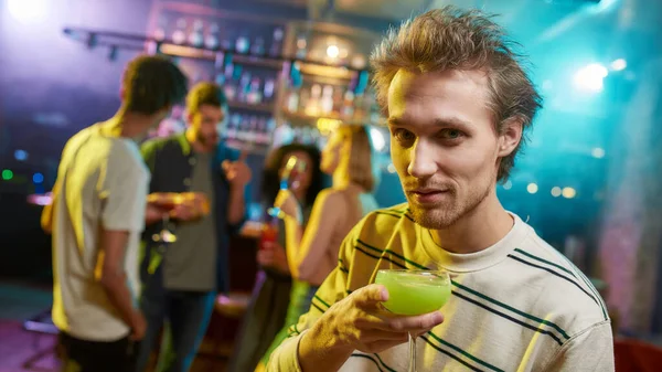 Ritratto di giovane caucasico che guarda la telecamera mentre posa con un cocktail in mano e gli amici che chiacchierano, bevendo al bancone del bar sullo sfondo — Foto Stock