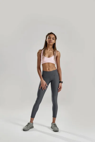 Foto de cuerpo entero de una joven mujer de raza mixta deportiva en ropa deportiva mirando a la cámara mientras posa aislada sobre un fondo gris. Estilo de vida saludable y concepto deportivo —  Fotos de Stock