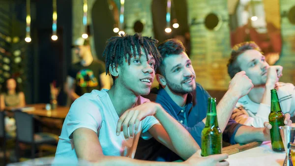 Cerveza para buenos amigos, tres hombres jóvenes bebiendo cerveza mientras ven el partido, amigos pasando tiempo en el bar deportivo, entusiasmados con el juego — Foto de Stock