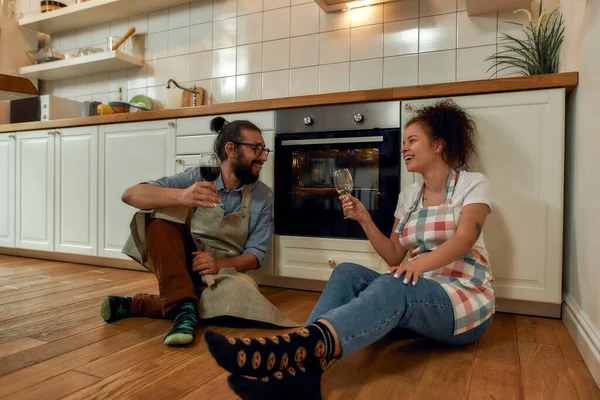 Fiatal boldog pár iszik egy pohár bort főzés közben a konyhában. Kötényes férfi és nő várja, hogy megfőzzön egy pizzát. — Stock Fotó