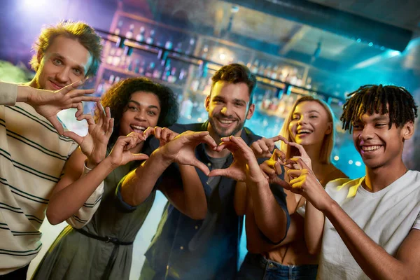Fiatal férfiak és nők, akik szívjeleket mutatnak, miközben együtt pózolnak a kamera előtt, multiracionális baráti társaság, akik a bárban buliznak — Stock Fotó