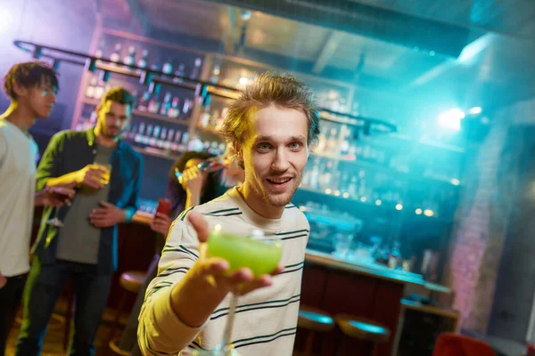 Caucásico joven mirando a la cámara mientras posando con un cóctel en la mano y amigos charlando, tomando bebidas en el mostrador del bar en el fondo — Foto de Stock