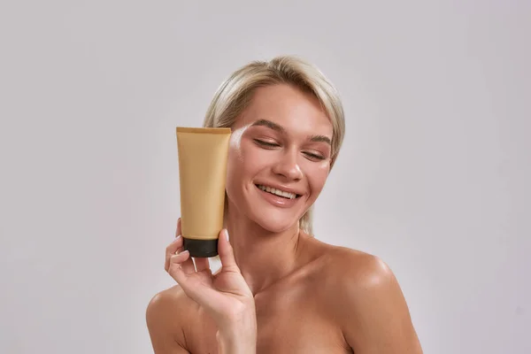 Portrait of young woman with perfect glowing skin smiling aside, holding sunscreen skincare product while posing isolated over grey background — Stock Photo, Image