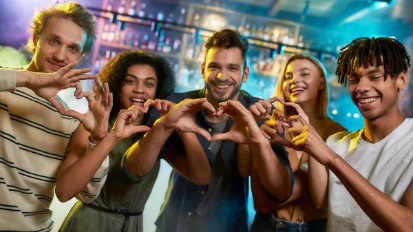 Eğlenceli bir geceydi. Genç erkekler ve kadınlar kameraya poz verirken kalp işaretleri gösteriyorlar. Çok ırklı bir arkadaş grubu barda takılıyor. — Stok fotoğraf