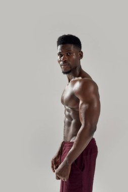 Genç kaslı Afro-Amerikan erkek çıplak gövdesiyle kameraya bakarken gri arka planda çıplak poz veriyor. Spor, spor, vücut geliştirme konsepti