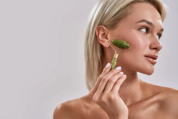 Close up portrait of young female blonde with perfect glowing skin looking aside while using jade facial roller for skincare and beauty treatment, posing isolated over grey background — Stock Photo, Image