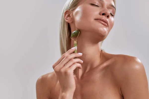 Retrato recortado de mujer joven con piel brillante perfecta usando rodillo facial de jade para el cuidado de la piel y el tratamiento de belleza mientras posa aislado sobre fondo gris — Foto de Stock