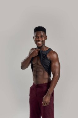 Atletik vücut. Genç kaslı Afrikalı Amerikalı adam kameraya gülümsüyor, siyah gömleğini kaldırıyor, karın kaslarını gösteriyor ve gri arka planda yalnız poz veriyor. Spor, spor, vücut geliştirme konsepti