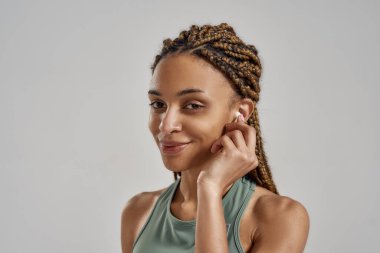 Antrenmandan sonra dinlenen, kablosuz kulaklık takan ve gri arka planda izole halde dururken müzik dinleyen mutlu, karışık ırk fitness kadını.