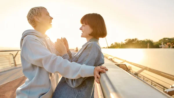 Lesbian couple looking at each other, laughing together while leaning on the bridge and watching the sunrise — Stock Photo, Image