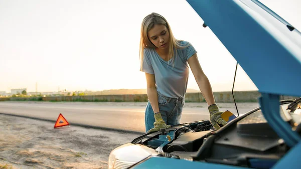 Zblízka záběr atraktivní mladá žena dívá pod kapotu svého rozbitého auta, snaží se opravit na vlastní pěst, zatímco stojí sám po autonehodě na silnici — Stock fotografie