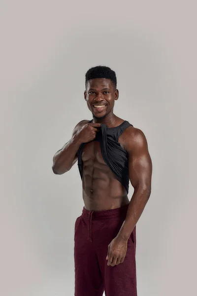 Corpo atlético. Jovem musculoso afro-americano sorrindo para a câmera, levantando camisa preta, mostrando seus abdominais enquanto posava isolado sobre fundo cinza. Esportes, treino, conceito de musculação — Fotografia de Stock