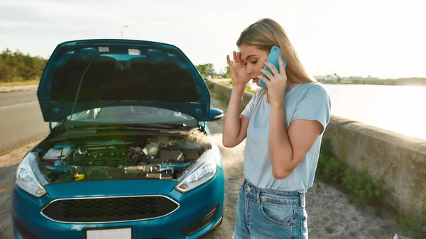 Aantrekkelijk meisje met problemen met de auto, Jonge vrouw met behulp van smartphone, bellen voor hulp of sleepwagen terwijl ze in de buurt van haar kapotte auto met open motorkap aan de kant van de weg — Stockfoto