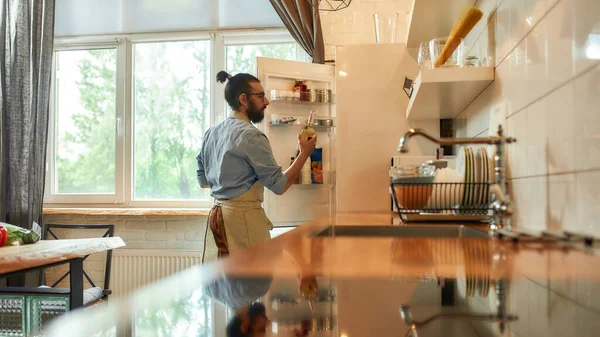 若い男,プロの料理人は、食事を準備しながら、冷蔵庫からワインや他の成分のボトルを取ってエプロン,キッチンに立って.家庭料理の概念 — ストック写真