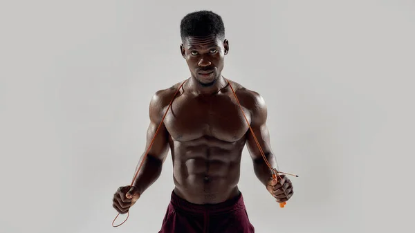 Homem americano africano jovem muscular olhando para a câmera, enquanto segurando corda de salto isolado sobre fundo cinza. Esportes, treino, conceito de musculação — Fotografia de Stock
