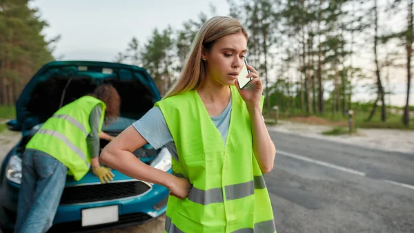 Paar dragen reflecterende vest hebben problemen met hun auto, Jonge vrouw bellen voor hulp of sleepwagen terwijl de man kijkt onder de motorkap van een gebroken auto — Stockfoto