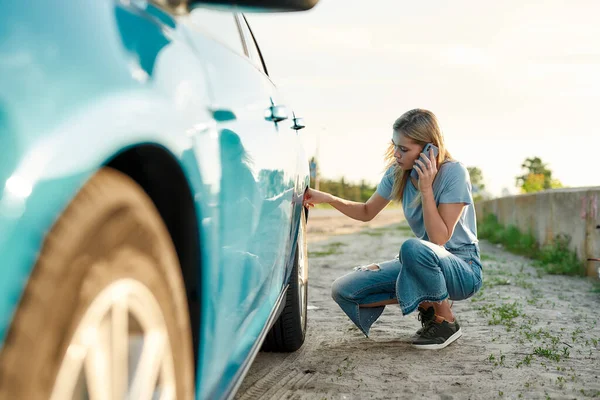 Aantrekkelijke jonge vrouw op zoek verdrietig, bellen auto service, bijstand of sleepwagen terwijl het hebben van problemen met haar auto, het controleren van wiel na auto pech op de lokale weg — Stockfoto