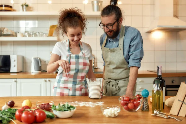 E 'ora della pizza. Giovane coppia innamorata di preparare l'impasto per fare la pizza con le verdure a casa. Uomo e donna indossano il grembiule, cucinano insieme in casa. Hobby, stile di vita — Foto Stock