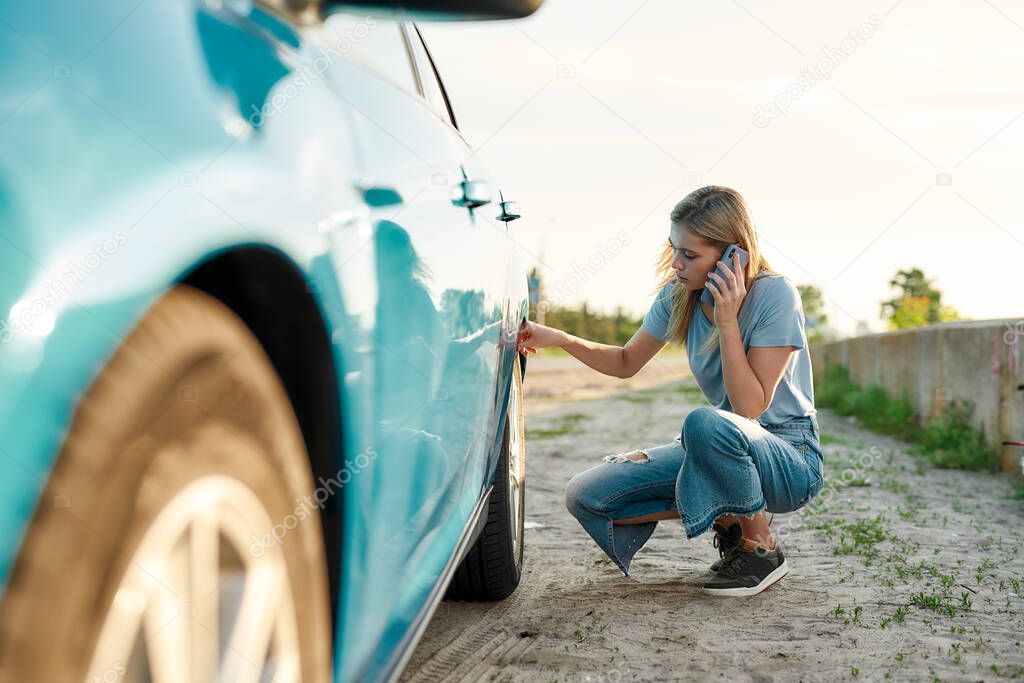 Attractive young woman looking sad, calling car service, assistance or tow truck while having troubles with her auto, checking wheel after car breakdown on local road side