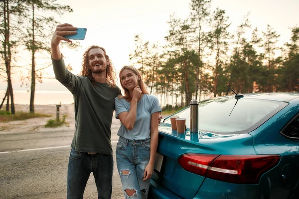 Homem alegre usando smartphone enquanto toma selfie junto com atraente jovem mulher depois de reparar carro quebrado no lado da estrada — Fotografia de Stock