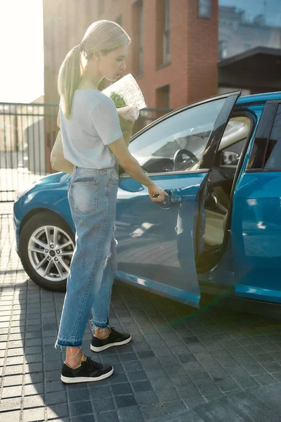 Full length shot van jonge vrouw openen van de blauwe auto deur terwijl het houden van boodschappentas na het winkelen in de supermarkt — Stockfoto