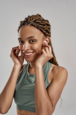 Genç ve güzel bir kadının dikey çekimi. Kablosuz kulaklık takan, müzik dinleyen, kameraya gülümseyen ve gri arka planda duran bir kadın.