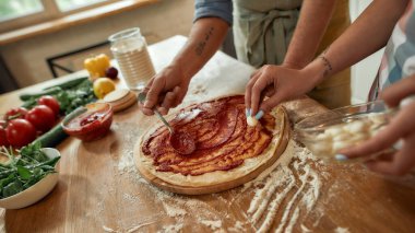 Takım çalışması. Evde birlikte pizza yapan bir çiftin görüntüsü. Önlüklü adam hamura domates sosu sürerken kadın mozzarella peyniri ekliyor. Hobi, yaşam tarzı