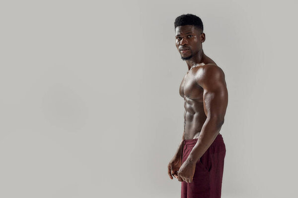 Чувствую себя хорошо. Молодой мускулистый африканский американец с обнаженным туловищем, смотрящий в камеру, позируя без рубашки на сером фоне. Спорт, тренировки, бодибилдинг