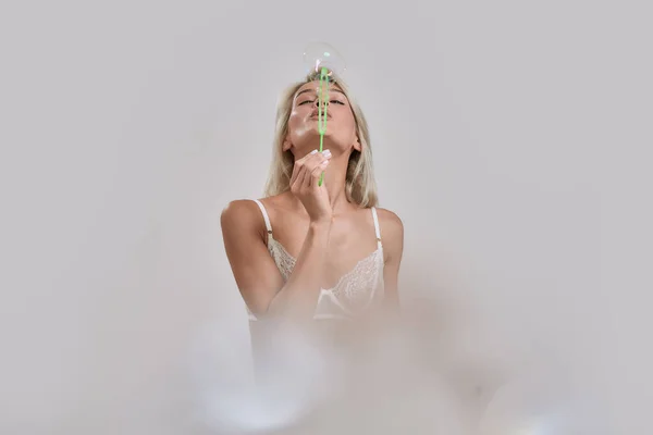 Sensuelle jeune femme en lingerie sexy s'amuser tout en soufflant des bulles de savon à la caméra, posant isolé sur fond gris — Photo