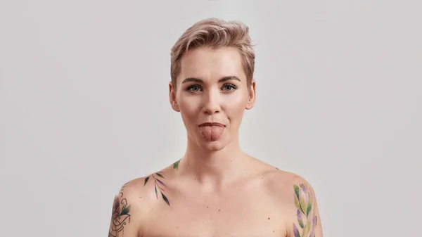 Retrato de uma jovem atraente mulher tatuada semi-nua com pele perfeita mostrando sua língua para câmera isolada sobre fundo claro — Fotografia de Stock