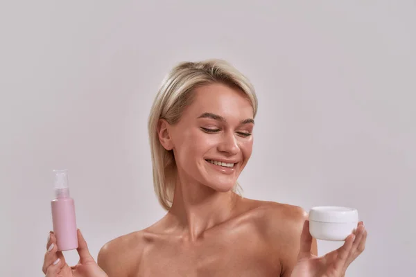 Porträtt av leende ung kvinna med perfekt hud hålla burk och flaska med olika kosmetiska hudvårdsprodukter och välja vad man ska tillämpa samtidigt posera isolerad över grå bakgrund — Stockfoto