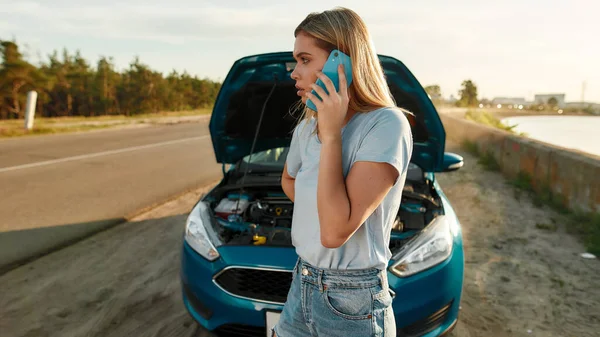 Aantrekkelijk meisje met problemen met de auto, jonge vrouw op zoek opzij en bellen voor hulp terwijl staan in de buurt van haar kapotte auto met open motorkap aan de kant van de weg — Stockfoto